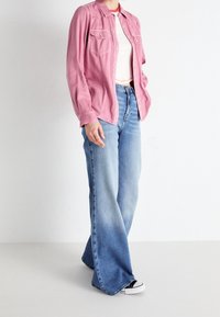 Chemise rose boutonnée avec deux poches poitrine, associée à un jean évasé bleu clair. Les chaussures présentent un design noir et blanc.