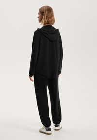 Črna kapuča s prostim krojem, v kombinaciji z črnimi joggers hlačami z elastičnimi manšetami. Bele superge z črnimi in sivo akcenti.