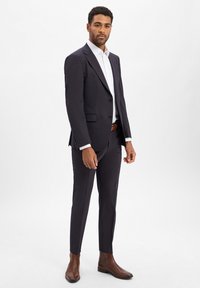 Marineblauw pak met een getailleerde pasvorm, een colbert met een enkele rij knopen en ingesneden revers, gecombineerd met een wit overhemd en bruine enkellaarsjes.