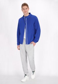 Giacca blu con zip e colletto rialzato, abbinata a pantaloni jogger grigi chiari con cordoncino e scarpe da ginnastica bianche. Tessuto morbido, vestibilità comoda.