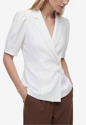 Vrouw draagt een witte korte mouwen blazer met opgeblazen schouders en twee knopen, gecombineerd met een bruine broek, handen in de zakken.