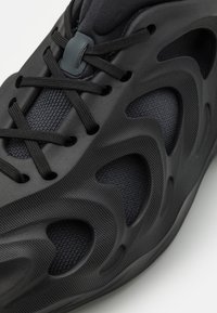 Svart atletisk sko med en texturerad ovandel, intrikat snörningssystem och utskärningar på sidorna för ventilation.