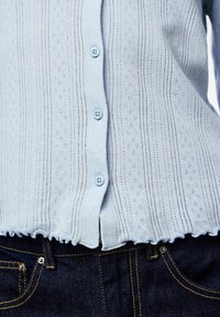 Ανοιχτό μπλε πλεκτό τοπ με κουμπιά και διάτρητο σχέδιο, με κυματιστό τελείωμα, συνδυασμένο με σκούρα μπλε τζιν παντελόνια με κεντήματα σε κίτρινο χρώμα για αντίθεση.