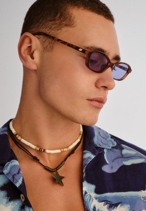 Homme portant des lunettes de soleil écaille de tortue, plusieurs colliers de perles et un pendentif en forme d'étoile, ainsi qu'une chemise bleue à motifs, regardant sur le côté.