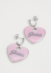 Orecchini a forma di cuore rosa con hardware in argento; presentano una finitura lucida e il logo grigio "Guess". Design con chiusura a perno.