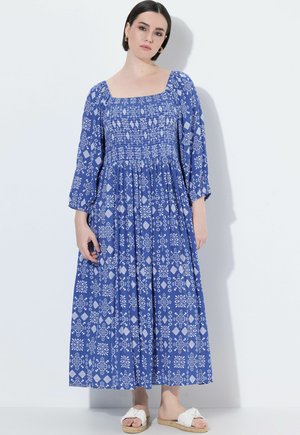 Kvinde iført en blå midi-kjole med hvide geometriske og blomsteragtige mønstre, firkantet udskæring, pufærmer til trekvartlængde og hvide slip-on sandaler.