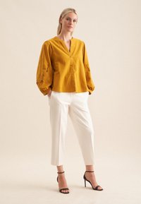 Camicia gialla senape con maniche a palloncino, che presenta dettagli ricamati, abbinata a pantaloni bianchi e sandali neri con tacco.