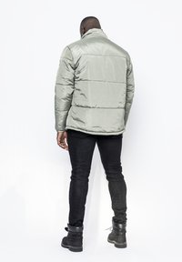Ljusgrön puffjacka med hög krage, med sydda paneler och en blank yta, som bärs över svarta jeans och robusta svarta stövlar.