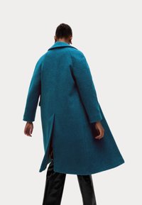 Abrigo extragrande color verde azulado hecho de tela texturizada, con cuello con solapa, mangas largas y una abertura en la parte trasera, combinado con pantalones negros.