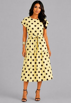 Femme portant une robe midi jaune à pois noirs, manches courtes, ceinture à la taille et talons noirs à bride cheville, debout devant un fond gris.