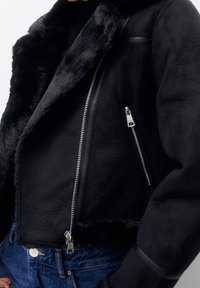 Chaqueta de piel sintética negra con un diseño cropped, que presenta un cierre con cremallera en la parte frontal, bolsillos laterales y detalles de cuero a lo largo de las costuras.