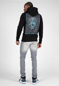 Sudadera negra con un gráfico turquesa en la parte trasera que presenta texto y un dibujo animado. Combinada con jeans gris claro y zapatillas deportivas oscuras.