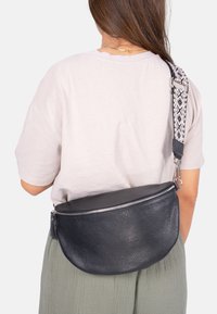 Schwarze Leder-Crossbody-Tasche mit strukturiertem Finish, silbernem Reißverschluss und gemustertem Tragegurt. Die Tasche hat eine runde Form und einen Reißverschlussverschluss.