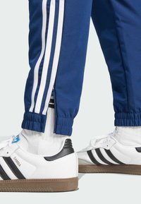 Białe sneakersy z czarnymi paskami, brązowe gumowe podeszwy, noszone z niebieskimi spodniami dresowymi z białymi bocznymi paskami i elastycznymi mankietami przy kostkach.
