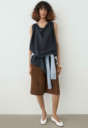 Kobieta z kręconymi włosami nosząca czarny, zdobiony drapowaniem top bez rękawów, brązową spódnicę do kolan, jasnoniebieski sweter zawiązany w talii, białe baleriny oraz złotą bransoletkę.