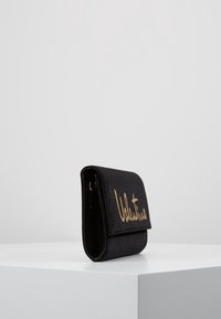 Svart tygplånbok med vikdesign, med en guld-signaturlogotyp på framsidan och lås med spänne. Slät textur.