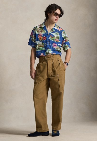 Jeune homme portant des lunettes de soleil, une chemise hawaïenne à manches courtes bleu à motifs, un pantalon marron taille haute avec de grandes poches, et des mocassins foncés.