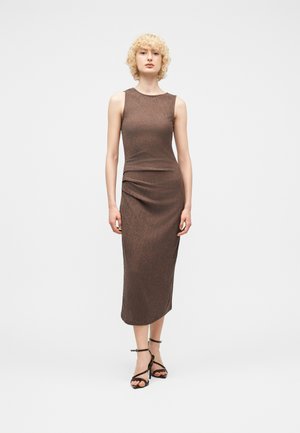 Moteris su garbanotais blondinų plaukais, vilkinti rankovių neturinčią rudos spalvos midi suknelę ir juodus aukštakulnius su dirželiais, stovinti priešais paprastą baltą foną.