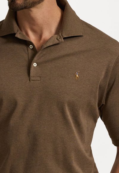 Rudas polo marškinėliai, pagaminti iš minkštos medžiagos, turi tris sagas, trumpas rankoves ir mažą išsiuvinėtą logotipą ant krūtinės.