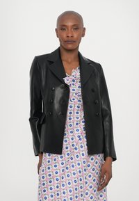 Čierny kožený blazer s dvojradovým zapínaním, ostrými klopami a priliehavými rukávmi, nosený cez šaty s kvetinovým vzorom.