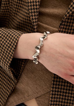 Bracciale in metallo argento con un design ondulato, caratterizzato da curve morbide e una finitura lucida, indossato su una mano contro un tessuto a motivo houndstooth.