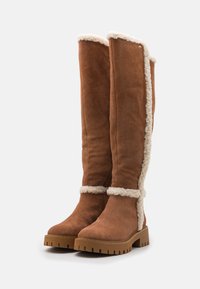 Brown suede knee-high boots with white faux fur lining, flat rubber sole, and chunky tread. Features a side seam accent with stitching details.