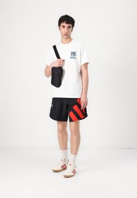 Fehér T-shirt kék grafikával, fekete rövidnadrág piros csíkokkal, fekete crossbody táska és bézs cipők piros akcentussal. Egyszerű háttér.