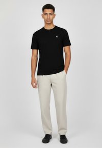 Czarny t-shirt z krótkim rękawem z małym logo na piersi, w połączeniu z jasnobeżowymi spodniami i czarnymi butami, stojący na szarym tle.
