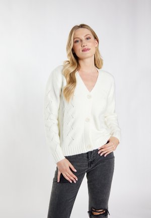 Witte gebreide cardigan met een V-hals, vijf knopen en een zigzagpatroon op de mouwen. Gecombineerd met versleten zwarte jeans.