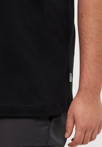 Tricou din bumbac negru cu tiv drept, mâneci scurte și un mic etichetă laterală. Textură netedă, design simplu și croială lejeră.