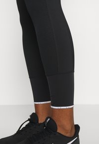 Czarne legginsy sportowe o dopasowanym kroju, z teksturowanymi panelami materiałowymi i białą logo na kostce. Noszone z czarnymi sneakerami.