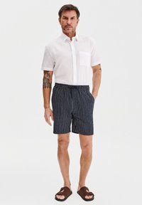 Biała koszula na krótki rękaw zapinana na guziki, granatowe spodenki w paski z elastycznym pasem, brązowe sandały, tatuaże widoczne na przedramieniu.