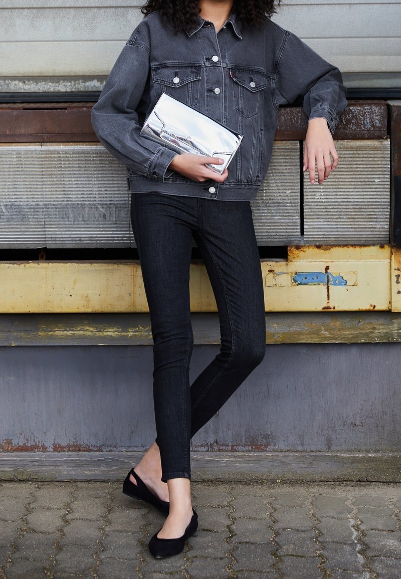 Grå denimjacka med bröstfickor, svarta skinny-jeans och svarta baletttofflor, håller en glänsande silverclutch mot en texturerad bakgrund.