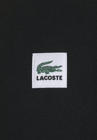 Étiquette en tissu blanc sur tissu noir texturé affichant un crocodile vert brodé au-dessus du texte « LACOSTE » en lettres capitales noires.