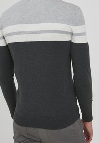 Mann in einem langärmligen Pullover mit grau, weiß und dunkelgrau horizontalen Streifen, kombiniert mit grauen Hosen, von hinten gezeigt.