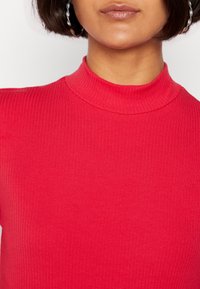 Femme portant un haut rouge à col montant côtelé avec des boucles d'oreilles en forme de cerceau tordu argenté, visible des épaules aux lèvres.