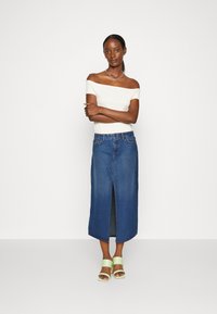 Jupe maxi en denim avec une fente avant, associée à un haut ajusté blanc cassé. Présente un lavage naturel et des détails de couture subtils. Sandales à talons vertes.