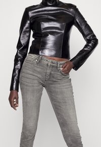 Chaqueta corta de cuero negro con cuello alto, textura suave y diseño ajustado, combinada con jeans grises de cintura media con un ligero desvanecimiento.