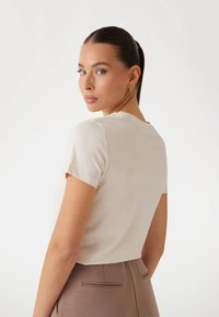 Maglietta beige chiara a maniche corte con scollatura tonda e texture morbida, abbinata a pantaloni taupe con tasche posteriori.