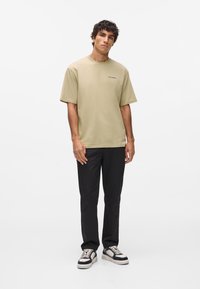 Olivovo zelené tričko s krátkym rukávom a okrúhlym výstrihom, s malým brandingom na hrudi, v kombinácii s čiernymi rovnými nohavicami a teniskami.