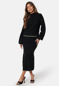 Zwart gebreid tweedelig outfit met ribbeltextuur, hoge halslijn en een aansluitende rok met subtiele accentdetails aan de zoom.