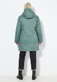 Giacca lunga, verde scuro, impermeabile, con cappuccio, caratterizzata da una texture liscia e un design minimalista, abbinata a jeans scuri e stivali neri.