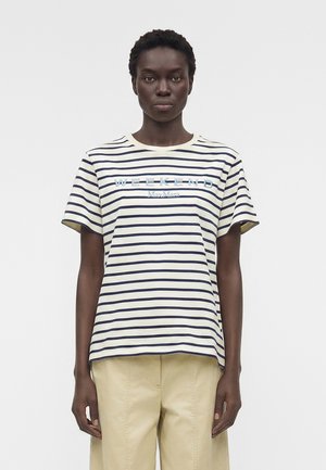 Γυναίκα που φοράει ένα μπλουζάκι με κοντά μανίκια σε λευκό και ναυτικό ριγέ χρώμα με το κείμενο "WEEKEND Max Mara", συνδυασμένο με μπεζ παντελόνι, στέκεται μπροστά σε απλό φόντο.