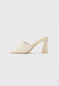Beige slide sandal with a textured upper adorned by white pearl accents. Features a geometric high heel for added support and style.