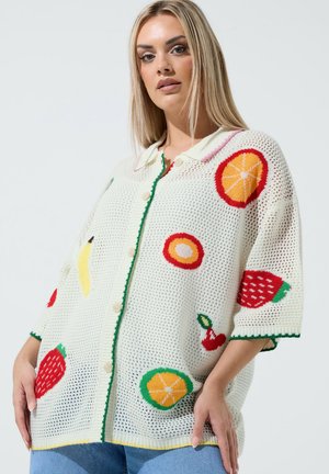 Blonde woman wearing a white knit cardigan with colorful fruit patterns and blue jeans against a plain gray background.