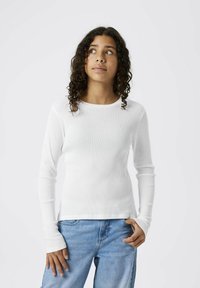 Weißes langärmeliges Rippoberteil mit Rundhalsausschnitt und figurbetonter Silhouette, kombiniert mit hellblauen Jeans. Glatte Textur und minimalistisches Design.