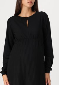 Schwarzes, geripptes Kleid mit rundem Ausschnitt und Schlüssellochdetail, das lange Ärmel und ein tailliertes Oberteil aufweist, das in einen lockeren Rock übergeht.