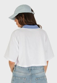 T-shirt bianca cropped con collo a righe blu, vista da dietro. Accompagnata da un berretto da baseball blu chiaro. Visibili sotto ci sono dei pantaloni denim.