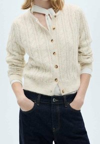 Cárdigan crema de punto con patrón de cables, escote redondo, botones de madera y puños acanalados, combinado con vaqueros azul oscuro.