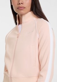 Lichtroze ritsjack met een hoge kraag, witte zijstrepen en een voorzak. Kenmerkt zich door gladde stof met minimale branding.
